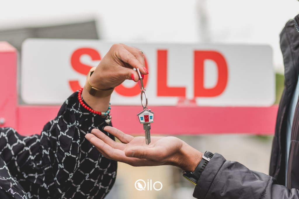 A seller handing the keys to a buyer in front of a sold sign.